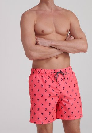 Mężczyzna bez koszulki, ze skrzyżowanymi ramionami, ubrany w koralowe spodenki kąpielowe z czarnym wzorem palmy i sznurkiem w talii, na jednolitym tle.