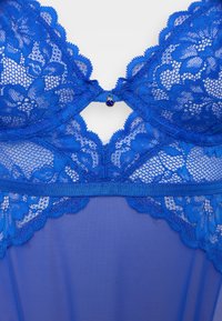 Lingerie blu in pizzo con motivi floreali, centro in rete trasparente, bordi scallop e un piccolo gioiello decorativo al centro.