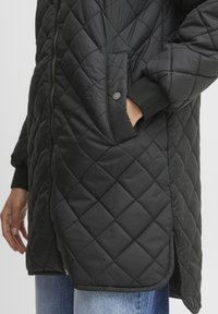 Schwarzer, gesteppter langer Mantel mit einem Reißverschluss, seitlichen Taschen und gerippten Bündchen. Das Material wirkt glatt mit einem Rautenmuster und abgerundetem Saum.