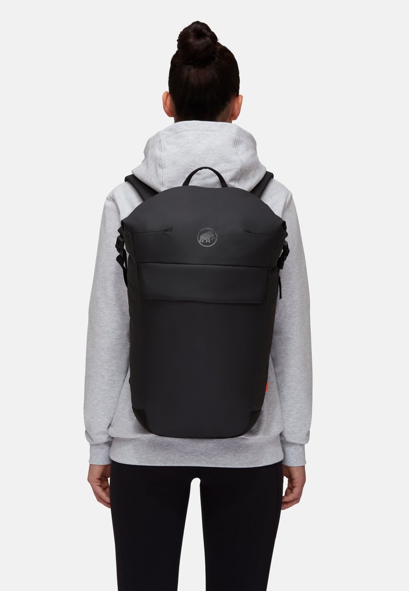Zwarte rugzak met een gladde, matte afwerking, afgeronde bovenkant, vo pockets en logo-accent; gedragen over een grijze hoodie met koordjes.