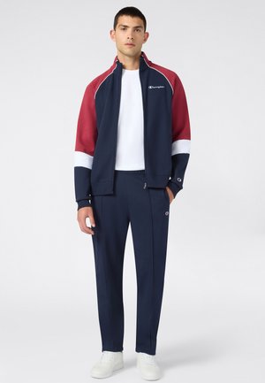 Jeune homme portant une veste de sport zippée bleu marine, rouge et blanche, un pantalon bleu marine assorti et des baskets blanches, debout devant un fond blanc uni.
