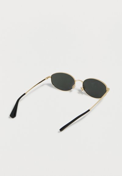 Γυαλιά ηλίου με χρυσή κορνίζα, οβάλ σκουρόχρωμους φακούς και μαύρες καμπυλωτές άκρες στους βραχίονες, τοποθετημένα σε λευκή επιφάνεια.