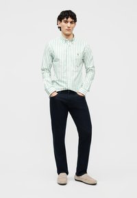 Άνδρας που φοράει πράσινο και λευκό ριγέ πουκάμισο με κουμπιά, σκούρο παντελόνι, μπεζ μοκασίνια, στέκεται με τα χέρια στις τσέπες μπροστά σε λευκό φόντο.