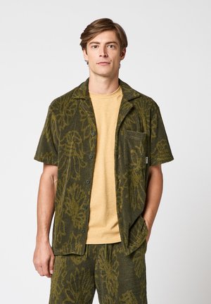 Jeune homme portant une chemise à manches courtes boutonnée vert olive et un short assorti avec un motif de feuilles, sur un t-shirt beige uni.