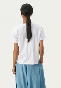 T-shirt bianca in cotone con maniche corte e taglio dritto. La parte posteriore presenta un semplice scollo e una texture liscia. Abbinata a pantaloni azzurri chiari.