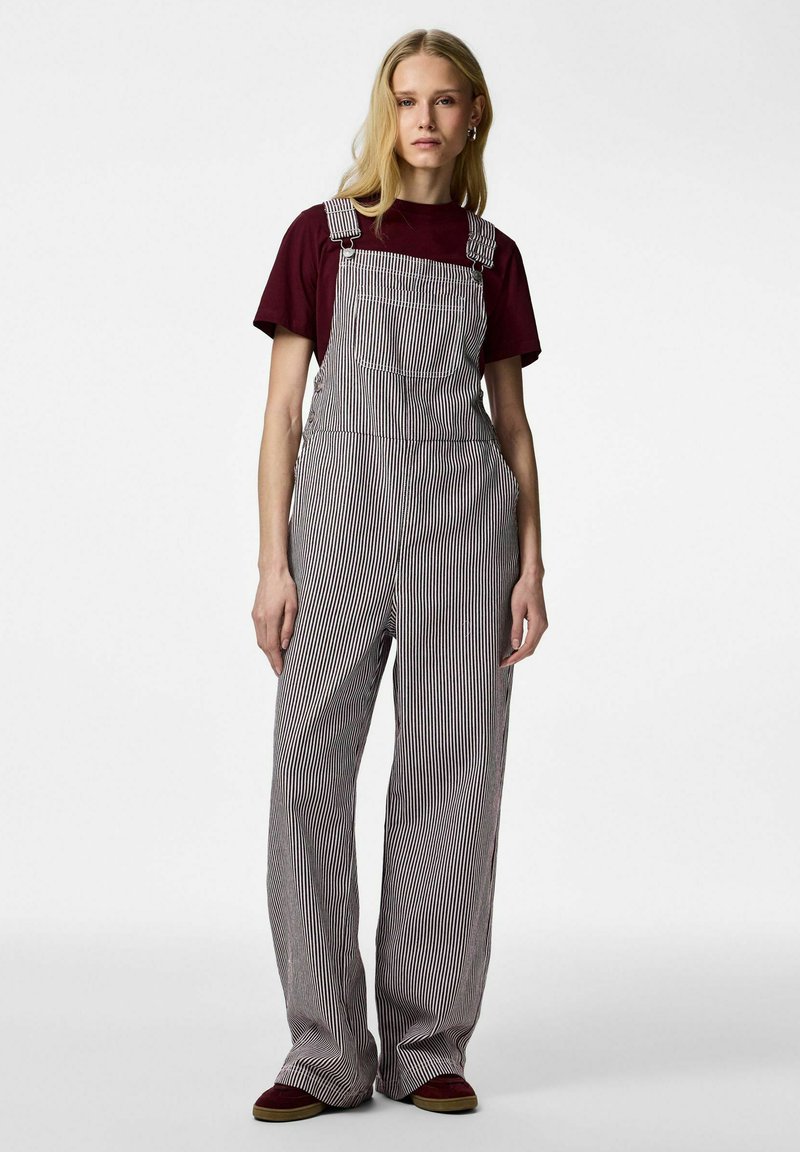 Blondínka stojaca, oblečená v čierno-bielych vertikálne pruhovaných nohaviciach na traky cez bordové tričko a zladené bordové topánky.