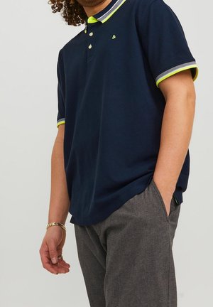 Mężczyzna w granatowej koszulce polo z neonowozielonym i białym wykończeniem, szarych teksturowanych spodniach, złotej bransoletce i pierścionku, z ręką w kieszeni.