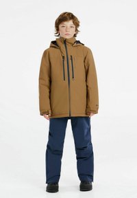 Braune wasserdichte Jacke mit zwei Reißverschlusstaschen, kombiniert mit marineblauen wasserdichten Hosen. Schwarze Stiefel, neutraler Hintergrund, aufrechte Haltung.