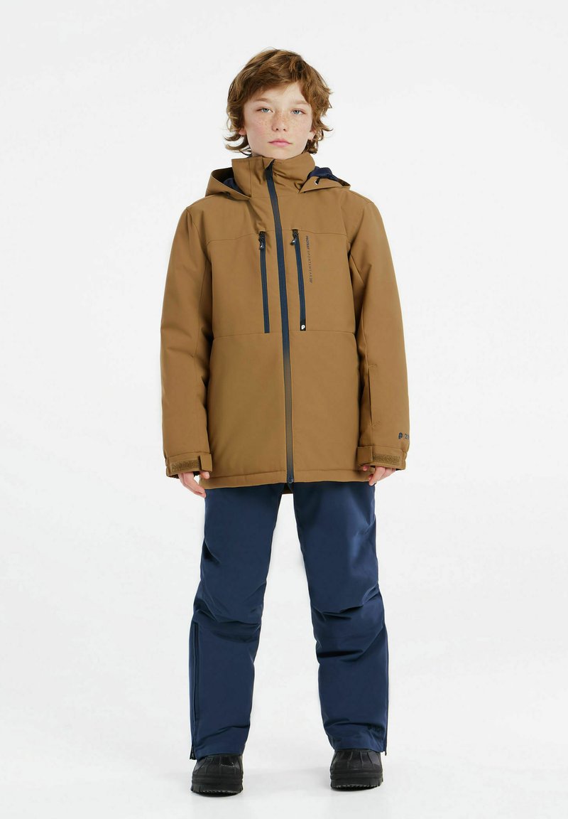 Braune wasserdichte Jacke mit zwei Reißverschlusstaschen, kombiniert mit marineblauen wasserdichten Hosen. Schwarze Stiefel, neutraler Hintergrund, aufrechte Haltung.