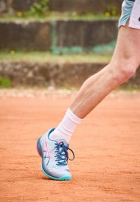 Baskets de sport bleu clair avec des accents roses et des lacets bleu foncé. Équipées d'un dessus texturé et d'une semelle en caoutchouc, posées sur un court de tennis en terre battue.
