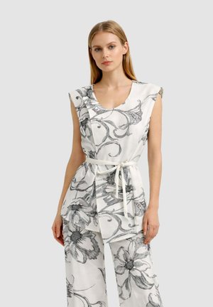 Donna che indossa una canotta bianca senza maniche e pantaloni con stampa floreale nera e cintura annodata, in piedi davanti a uno sfondo grigio semplice.