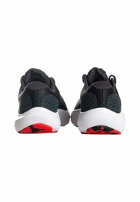 Scarpe atletiche con una tomaia in rete nera, dettagli grigi e un’intersuola bianca. La suola presenta un motivo rosso e nero con scanalature per trazione.