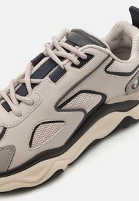 Sportos sneaker, amely bézs sima bőrből készült felsőrésszel rendelkezik, hálós panelek, sötét tengerészkék díszítések és vastag, texturált krémszínű gumitalppal.