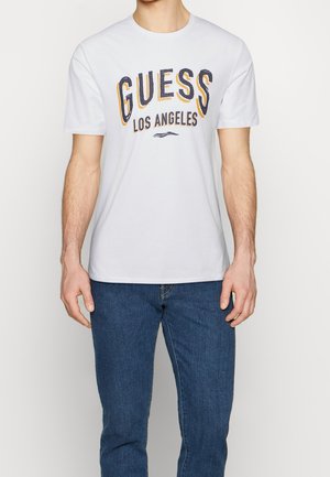 Mand iført hvid kortærmet T-shirt med teksten "GUESS Los Angeles" og blå jeans, stående foran en ensfarvet lys baggrund.