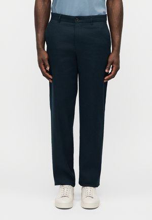 Mężczyzna ubrany w granatowe spodnie o prostych nogawkach, jasnoniebieską koszulę i białe sportowe buty, stojący z ramionami swobodnie opuszczonymi wzdłuż ciała.