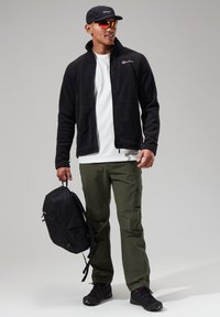 Black fleece zip jacket, white t-shirt, olive cargo pants, black backpack, black shoes. Model wearing cap and reflective sunglasses.
