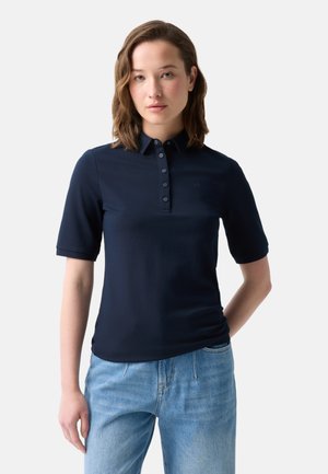 Jonge vrouw met kort golvend haar, gekleed in een donkerblauw poloshirt met korte mouwen en lichtblauwe jeans, staand tegen een effen achtergrond.