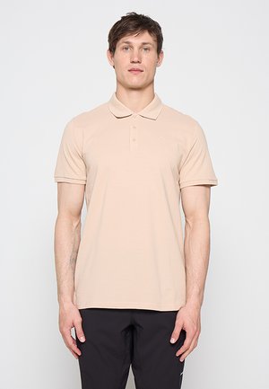 Jonge man die vooruit kijkt, draagt een beige polo met korte mouwen en een zwarte broek tegen een effen lichte achtergrond.