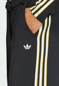 Svarta joggingbyxor med vita och gula sidostripes, försedd med en liten vit logotyp på fickan och texturerat tyg.