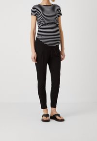 Umstandsmode mit einem schwarz-weiß gestreiften Oberteil mit Raffung und schwarzen, tapered Hosen mit Kordelzugbund. Schwarze Sandalen runden das Outfit ab.