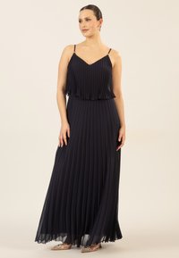 Marineblaues, gefaltetes Maxikleid mit dünnen Trägern und einem geschichteten Design. Leichter Stoff fällt sanft und zeichnet eine fließende Silhouette mit durchscheinenden Details.