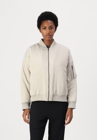 Lys beige bomberjakke med glidelås foran, ribbestrikkede mansjetter og kant. Har en sidelomme med glidelås og stående krage. Glatt tekstur.