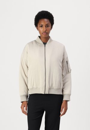 Lys beige bomberjakke med glidelås foran, ribbestrikkede mansjetter og kant. Har en sidelomme med glidelås og stående krage. Glatt tekstur.