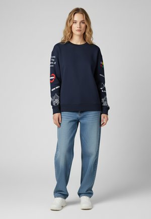 Felpa navy a maniche lunghe con diverse grafiche in bianco e rosso, abbinata a jeans in denim blu chiaro e sneakers bianche.