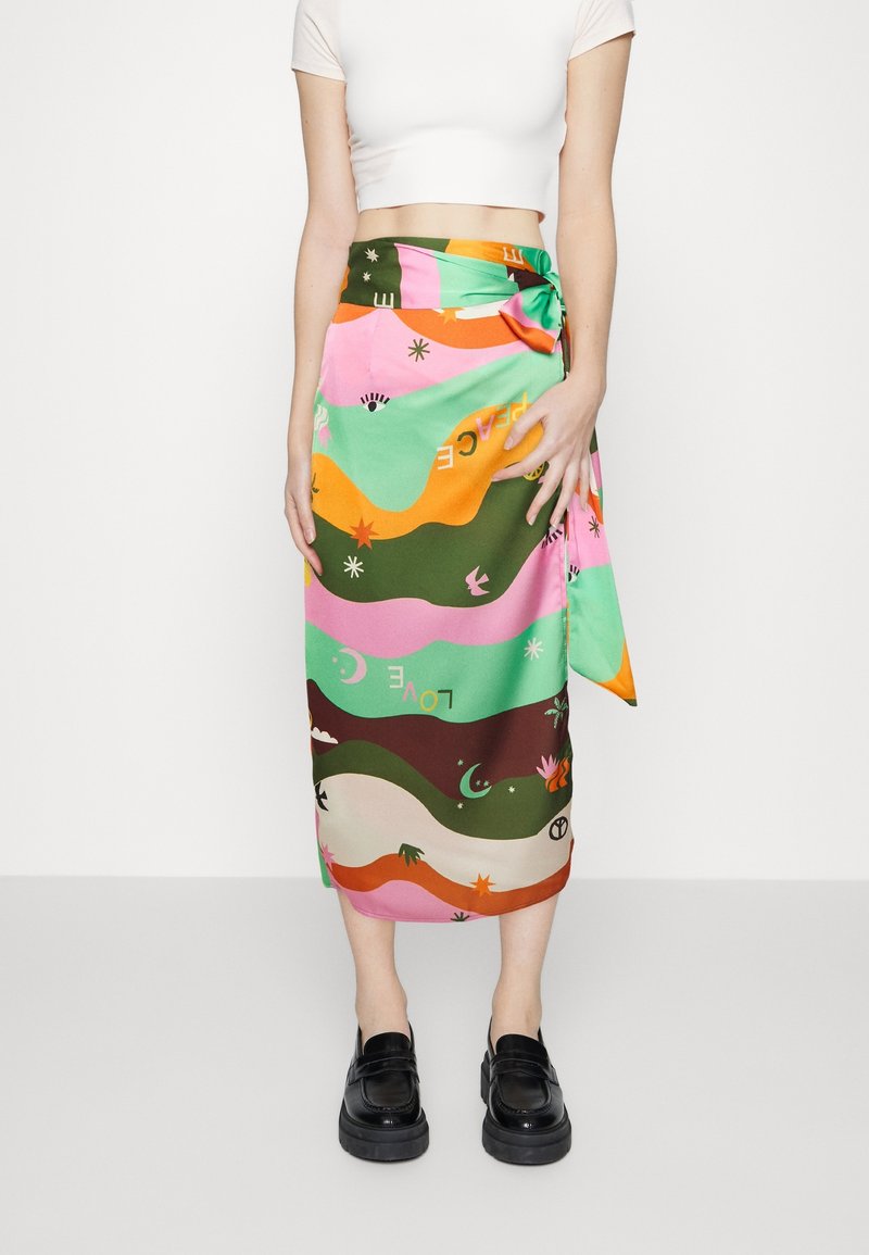 Femme portant une jupe portefeuille colorée avec des motifs abstraits et des mots, associée à un crop top blanc et des mocassins à plateformes noirs.