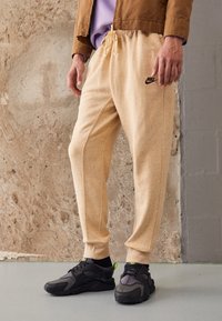 Vaaleanbeiget juoksuhousut, jotka on valmistettu teksturoidusta kankaasta ja joissa on kiristysnauha vyötäröllä. Sivusaumat ja musta Nike-logo. Yhdistetty mustiin tennareihin.