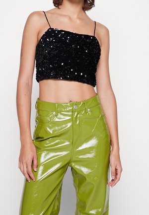 Kvinde iført en sort, palietbesat cropped tanktop og højtaljede, blanke limegrønne bukser mod en ensfarvet baggrund.