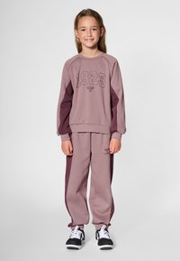 Lila sweatshirt med "1923"-tryck, kombinerad med matchande mjukisbyxor med vinröda sidopaneler, bärs med svarta sneakers.