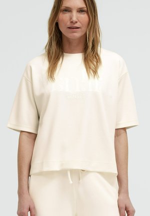 Woman with shoulder-length blonde hair wearing a loose off-white short-sleeve top and matching pants against a plain background.