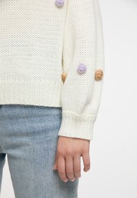 Fehér kötött pulóver texturált mintával, lila és bézs pom-pom díszítéssel az ujjakon. Világoskék farmernadrággal viselve.