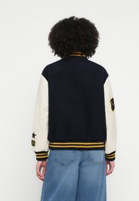 Tengerészkék egyetemi dzseki krémszínű ujjakkal, bordázott mandzsettákkal és szegéllyel. Sárga és fekete csíkos díszítésekkel és bal oldali ujján jelvénnyel rendelkezik.