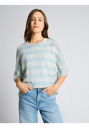 Junge Frau mit langen braunen Haaren, die ein locker gestricktes Top mit hellblau-weißen Streifen und hellblaue Jeans trägt, Hände in den Taschen, neutraler Hintergrund.