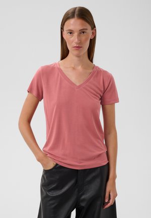 Femme aux cheveux bruns et raides porte un t-shirt rose poussiéreux à manches courtes et col en V, ainsi qu'un pantalon en cuir noir avec une main dans la poche.