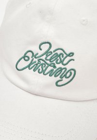 Casquette en coton blanche avec une visière courbée, portant un texte brodé vert indiquant "Just Exist". Le tissu a une finition texturée avec des détails de couture.