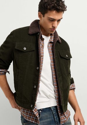 Jeune homme portant une veste en velours côtelé vert foncé avec un col en fourrure retournée, sur une chemise marron à carreaux et un t-shirt blanc, regardant vers le bas.