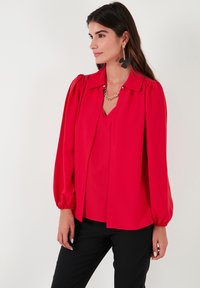 Red blouse with long, puffed sleeves and a V-neckline. Smooth texture, tailored fit, featuring a buttoned collar and subtle pleats.
