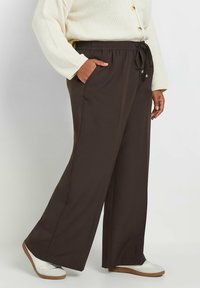 Bruine, wijde broek met een elastische tailleband, koordsluiting en zijzakken. Gecombineerd met een crèmekleurige trui en witte schoenen.