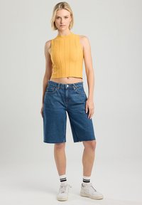 Keltainen ribattu toppi korkealla kauluksella yhdistettynä sinisiin farkku-bermudashortseihin, joissa on suora leikkaus ja riiputetut helmat. Valkoiset lenkkarit mustilla yksityiskohdilla.