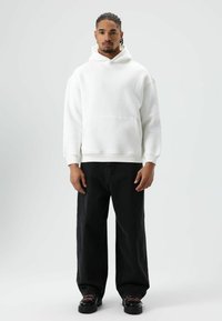 Witte sweater met capuchon en een voorzak, gecombineerd met losse zwarte jeans en zwarte schoenen met roze veters. Gladde textuur en ontspannen pasvorm.