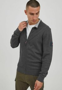 Dark grey zip-up cardigan with a light grey collar, ribbed cuffs, and an emblem on the left sleeve. Smooth fabric texture.
