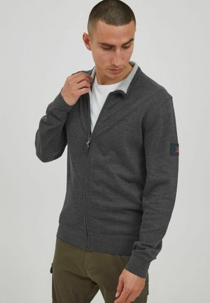 Cárdigan de cremallera en gris oscuro con un cuello gris claro, puños acanalados y un emblema en la manga izquierda. Textura de tela suave.
