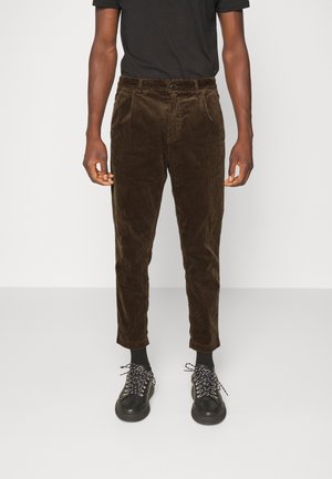 Mand iført brune kordbukser, sorte sokker, sorte sneakers med hvide snørebånd og en sort kortærmet skjorte, stående mod en hvid baggrund.