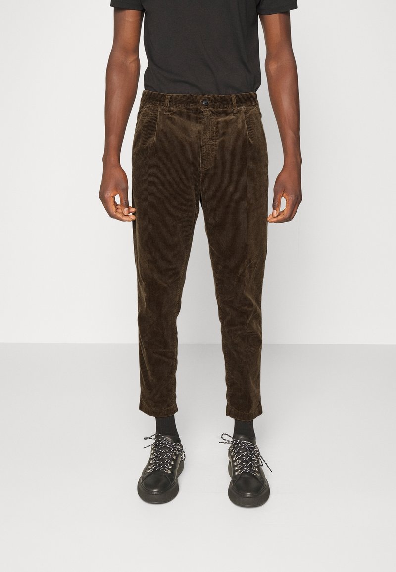 Homme portant un pantalon en velours côtelé marron, des chaussettes noires, des baskets noires avec des lacets blancs et une chemise noire à manches courtes, debout sur un fond blanc.