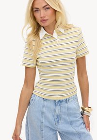 Femme aux longs cheveux blonds portant un polo à manches courtes rayé jaune, blanc et marron et un jean taille haute bleu clair.