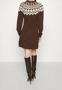 Robe en tricot marron avec un motif géométrique blanc au niveau des épaules, à manches longues, et une coupe décontractée, associée à des bottes hautes marron foncé.