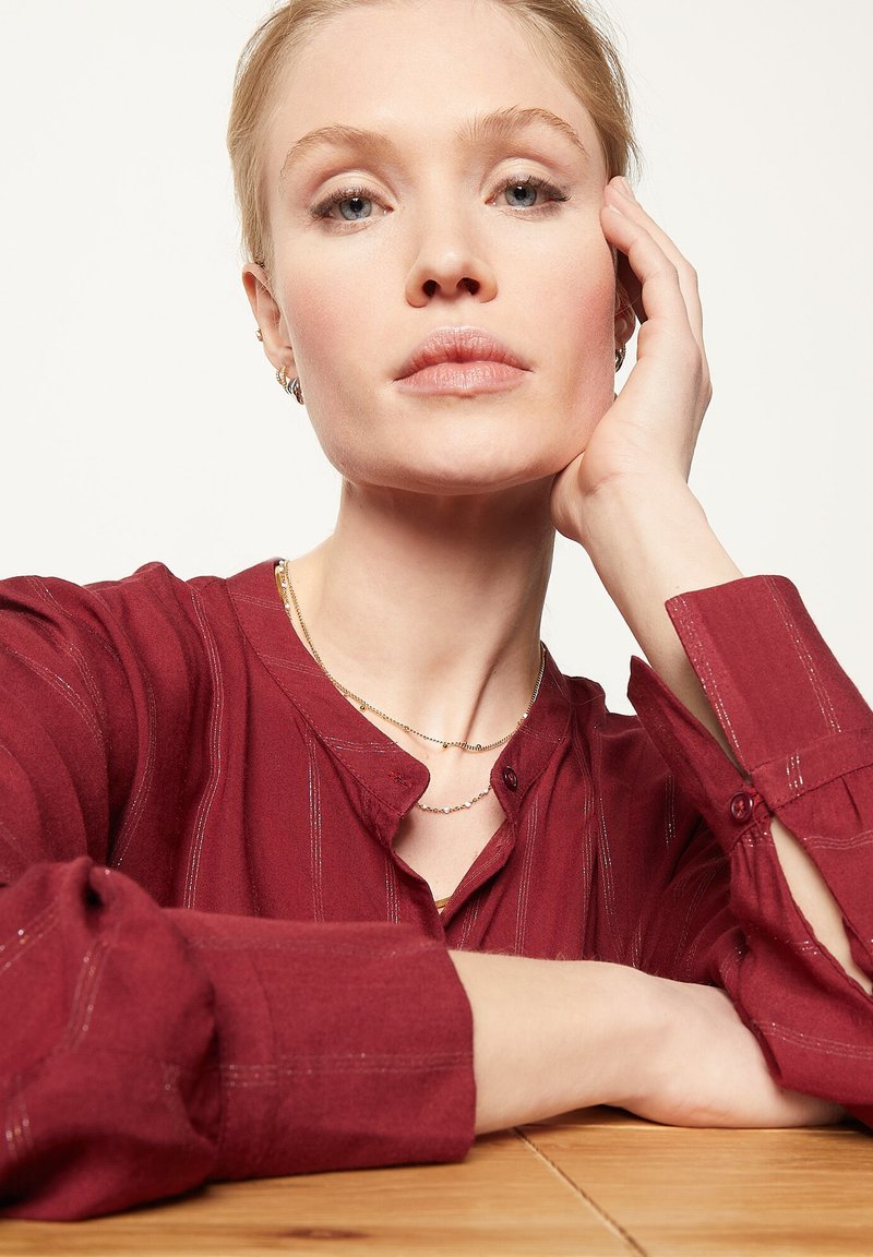 Junge Frau mit blonden Haaren stützt ihren Kopf auf ihre Hand, trägt ein rotes Langarmshirt und zwei zarte Halsketten über einem Holztisch.